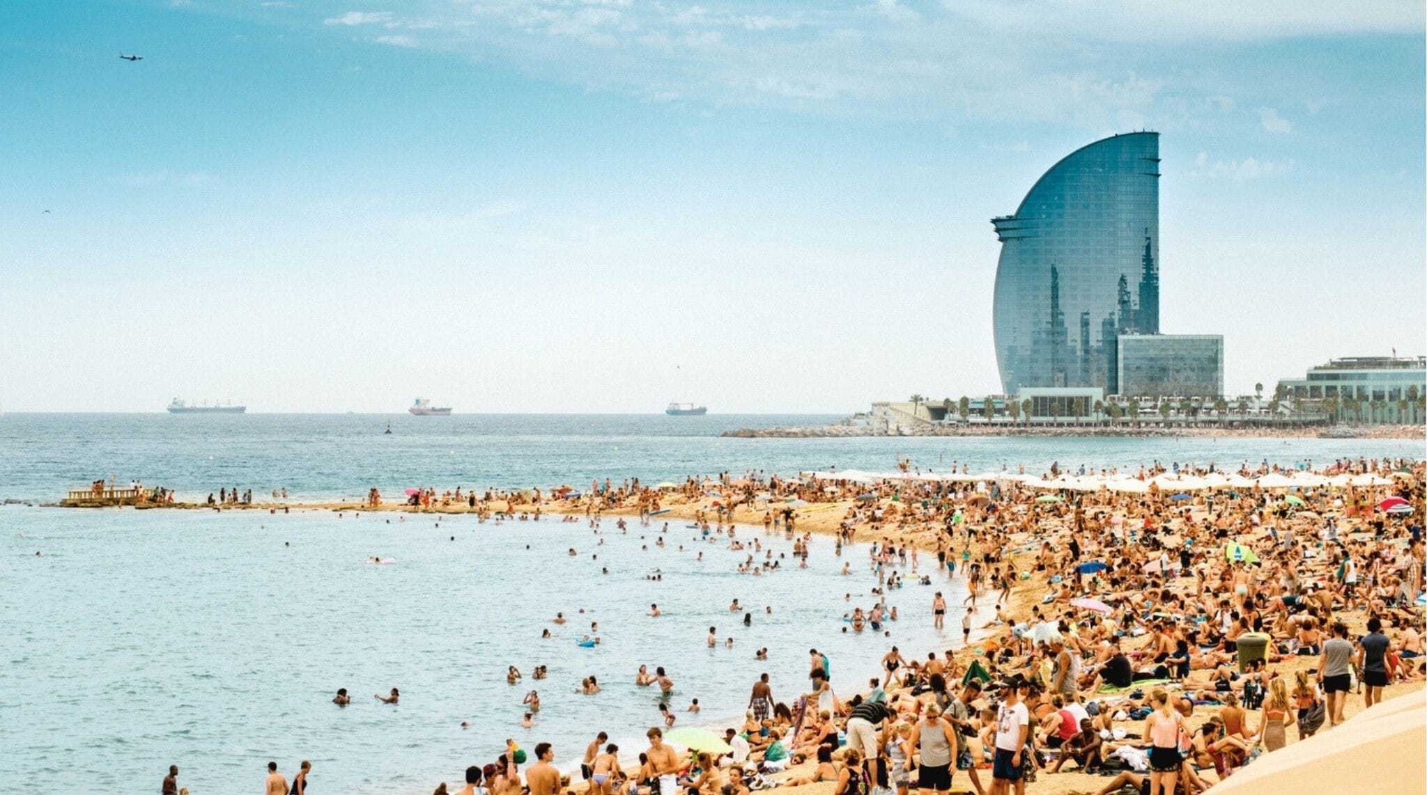 Playa de la Barceloneta llena de gente con el icónico hotel vela al fondo en Barcelona
