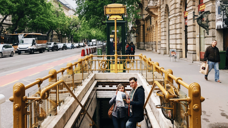 A photograph of the Foldalatti metro stop in Budapest. There is a sign with the metro stop's name on it in the background. In the foreground, there are some stairs leading down into the metro station. On the stairs, a man and woman have stopped and are looking at a map of Budapest. To illustrate a blog post entitled 'The Ultimate Sziget Festival Guide.'