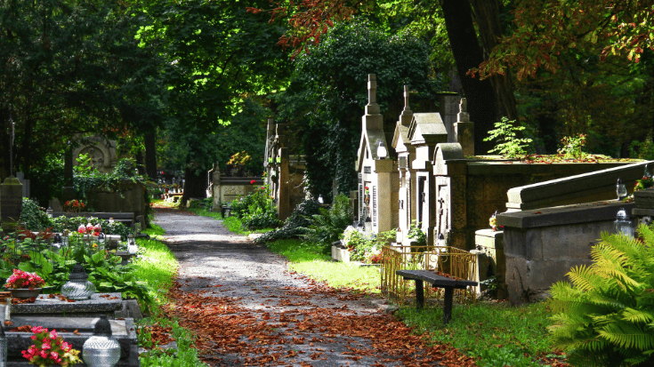 A colour photograph of a grey gravel path winding through a peaceful cemetery. There is light shining above from the sun, dappled by the leaves of overhanging trees. The leaves on the trees are green and there are lines of beautifully carved and well-kept sand-coloured tombstones along the path. To illustrate a blog post entitled '10 of The World's Most Beautiful Cemeteries'.