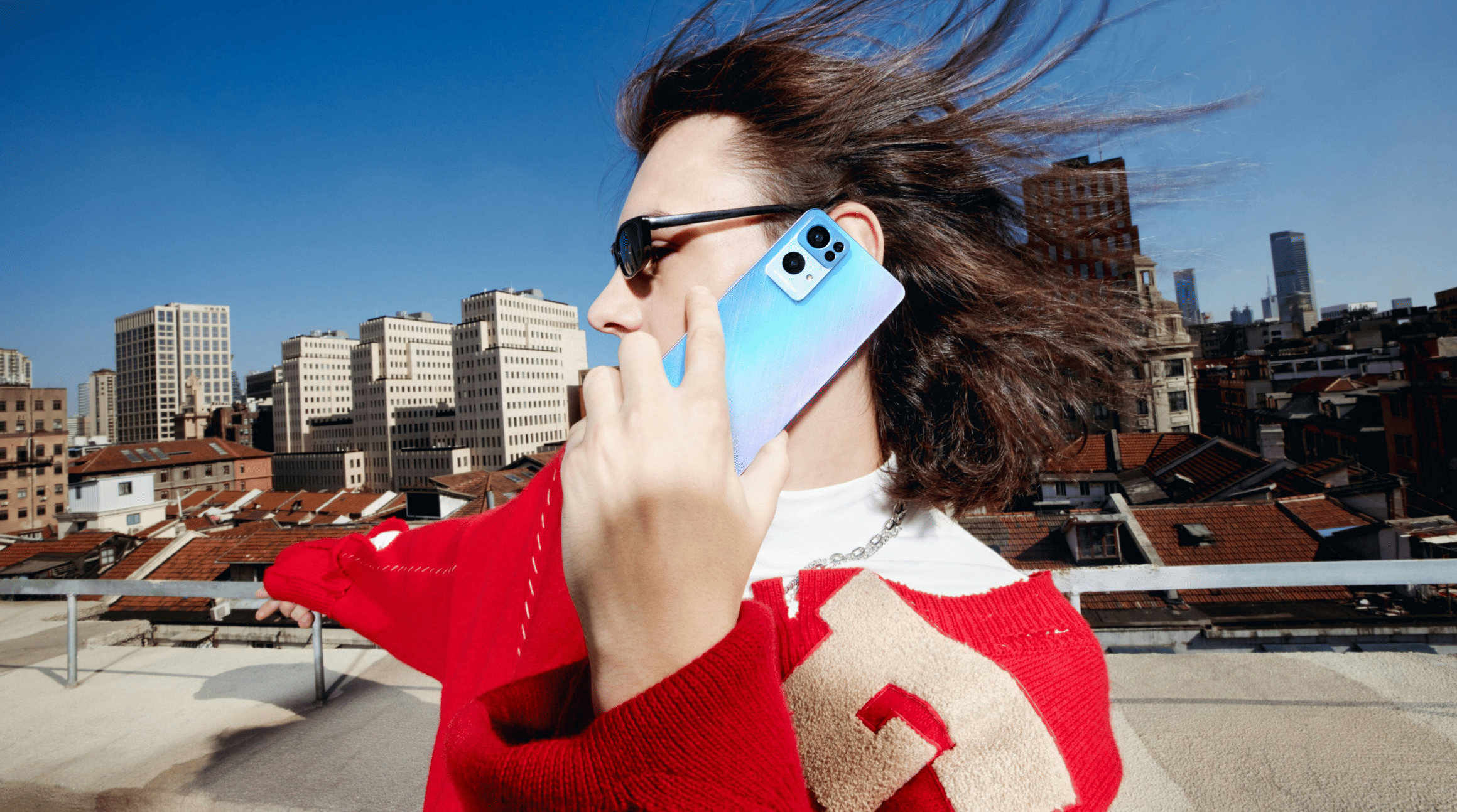 A colour photograph of a woman standing on a rooftop balcony. in the background, there are red slate rooftops and apartment blocks in colours such as white, orange and beige. She is wearing a red and white jumper, has brown hair and you can see her from a side profile. She is talking on an Oppo phone, with an eSIM from Airalo. To illustrate a blog post entitled 'How to Add an eSIM to an Oppo Phone.'