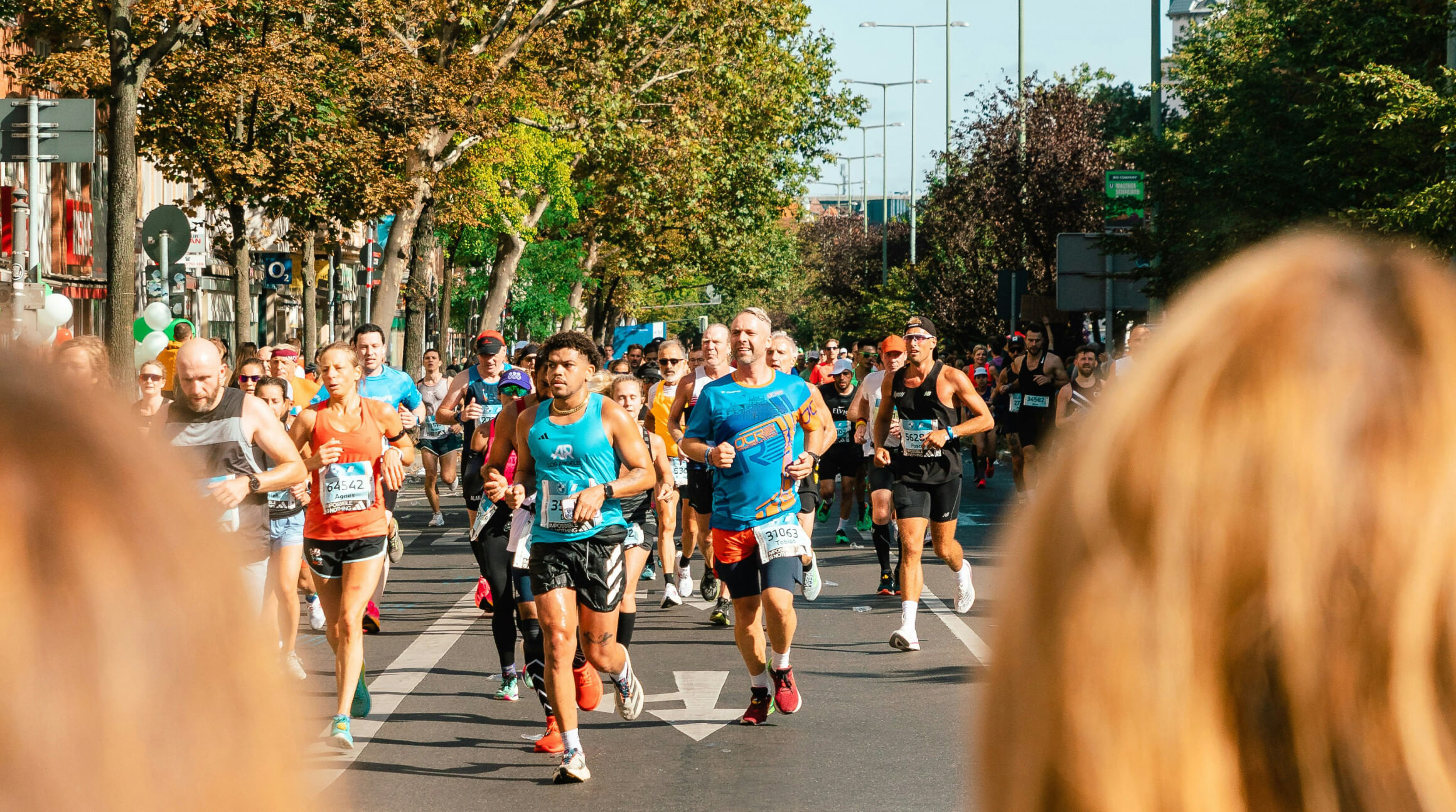 A colour photograph showing a large crowd of runners pacing the streets of Berlin. They are all wearing race numbers and different coloured running clothes, such as shorts, compression socks and lightweight t-shirts. In the background are trees and a clear blue sky. To illustrate a blog post entitled 'Berlin Half Marathon 2026: Can You Still Register?'