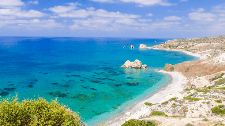 A photograph of a secluded white-sand beach winding up a coast in the southwest of Cyprus. The sky is bright blue with some white clouds, the sea is azure blue and there is some greenery along some sand dunes. In the water just off the coast is a beige rock, named the Rock of Aphrodite. To illustrate a blog post entitled 'Where is Hot in November in Europe?'