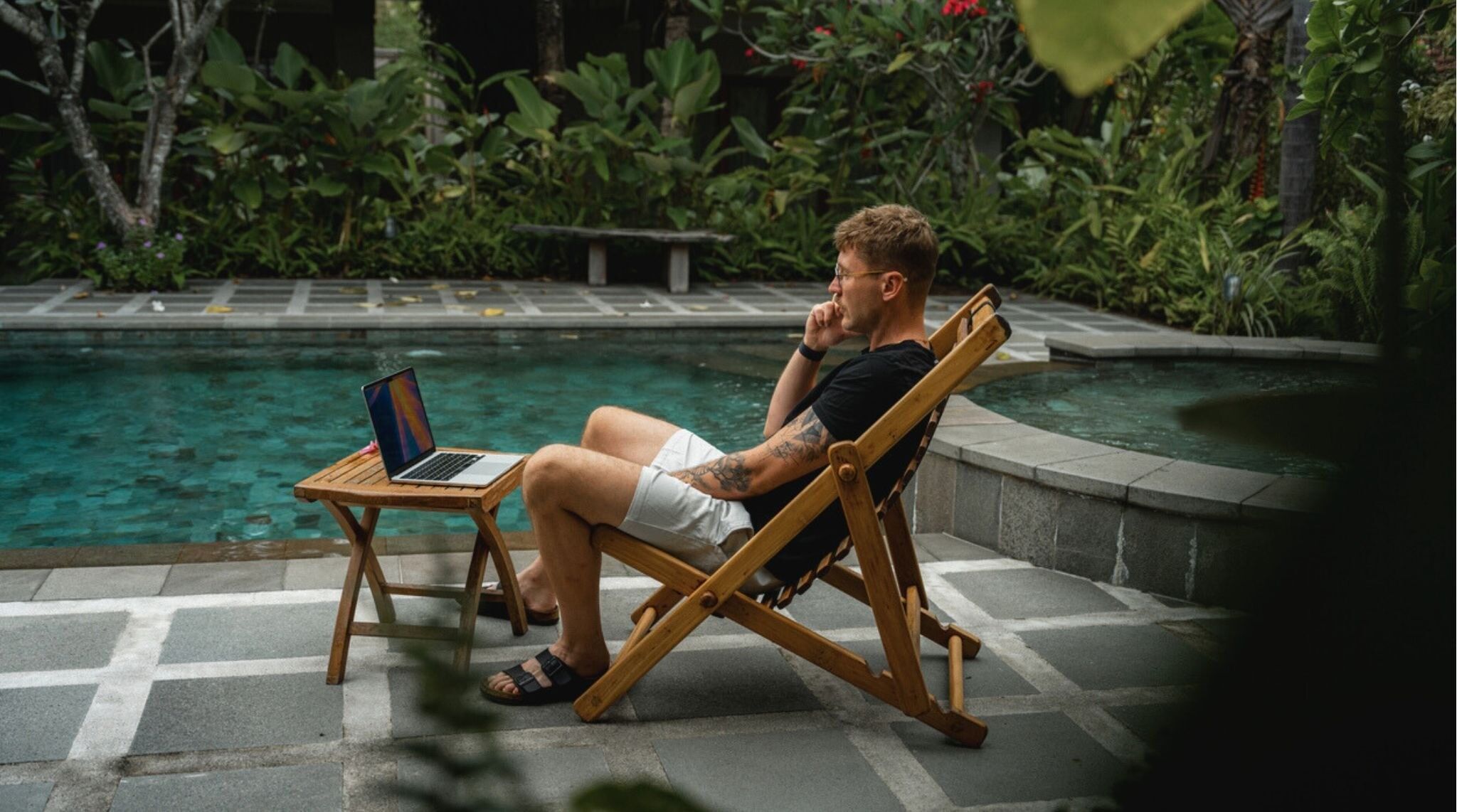 Homem falando ao telefone enquanto usa notebook, próximo a piscina em jardim tropical.