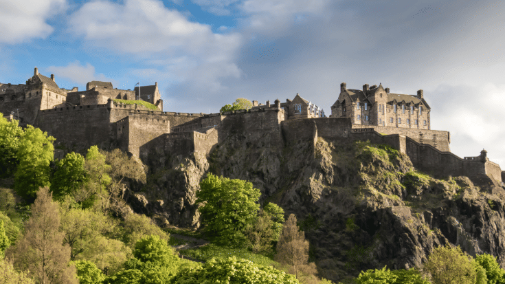 A colour photograph of Edinburgh Castle sitting on a red/brown cliff. It shows the building's walls and outhouses and underneath some greenery in the summer time, including trees and bushes. The sky in the background is a clear blue with some white clouds. To illustrate a blog post entitled 'Guillermo Del Toro's Frankenstein Filming Locations.'