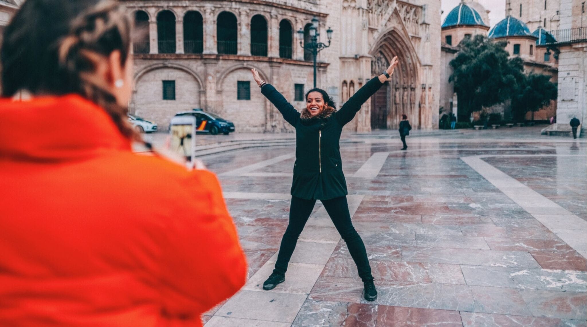 Uma turista registra um momento alegre, fotografando sua amiga com os braços abertos em frente à famosa Catedral de Valência durante um passeio pela cidade. O cenário de inverno, a arquitetura histórica e o gesto entusiasmado representam perfeitamente o espírito de viagem e exploração na Espanha.
