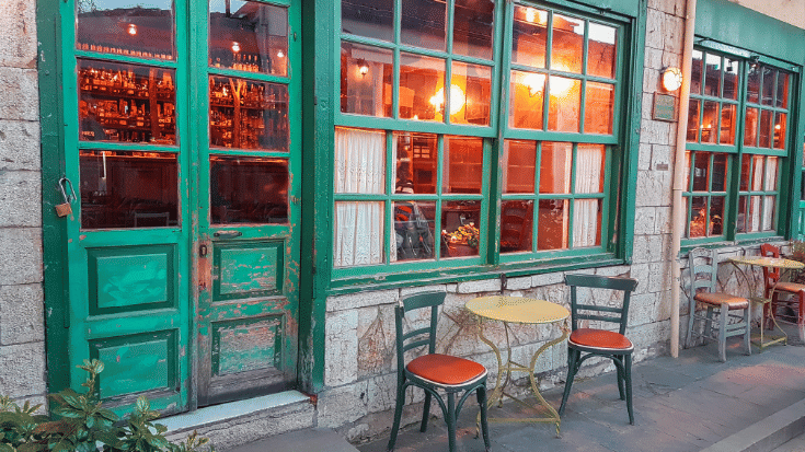A photograph of an old-school coffee shop in Greece. The window frames and door are covered in an emerald green pain tthat is peeling with age. There are glowing orange incandescent lights inside showing shelves and shelves of drinks, spirits and other bottles. Outside is a single table and two chairs, which are also green with red seating pads. The outside of the coffee shops has stone walls. To illustrate a blog post entitled 'Which Country Has the Best Coffee Culture?'