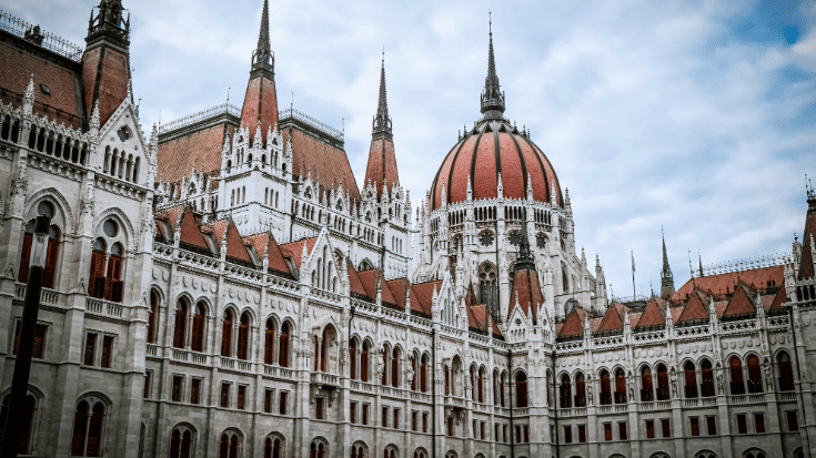A photograph of Budapest's large Gothic-style parliament building. It shows many windows and the decorative arches above them. The building is in a grey stone colour, while there are domes above the windows in a striking rust/red colour, all against a blue sky with white clouds. To illustrate a blog post entitled 'The Ultimate Sziget Festival Guide.'