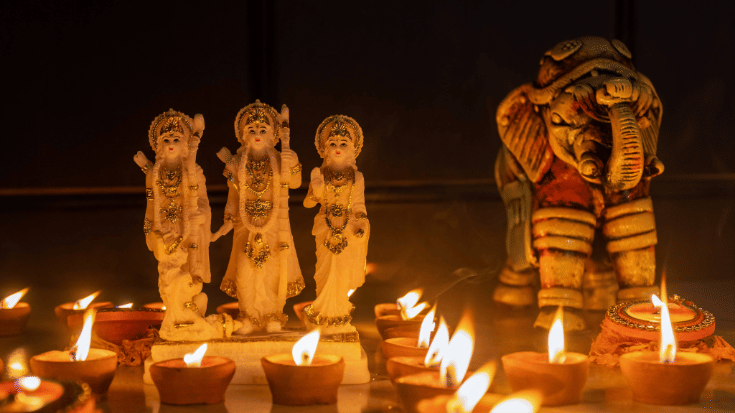 A photograph of a statue of Lord Rama, brightened by traditional Indian Diya candles for Diwali. The background is dark and the small stone statue is illuminated in the foreground by the lit candles with an orange glow. Diwali is the Hindu festival of lights, celebrated in India. To illustrate a blog post entitled 'When Is Diwali 2025? Your Questions Answered.'