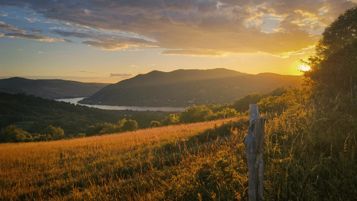 A landscape photograph of a countryside scene in Visegrad, Hungary. The sky is streaked with an orange and pink sunset, against a mountain skirted by a river. In the foreground is a golden field and a walk way. To illustrate a blog post entitled 'The Ultimate Sziget Festival Guide.'