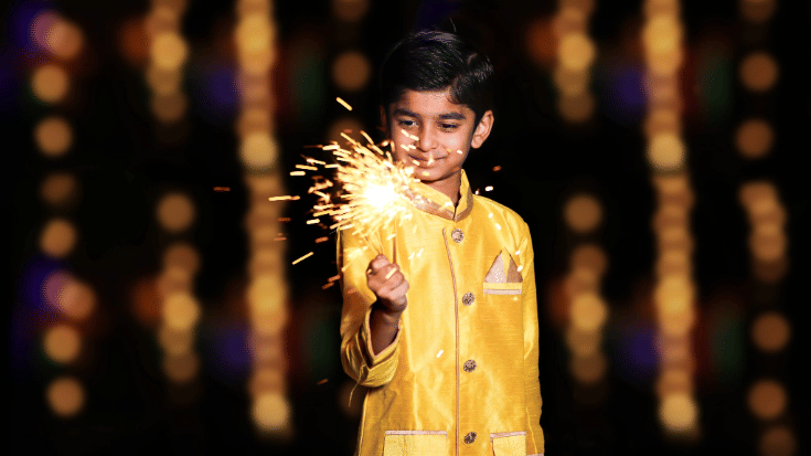 A photograph of a small Indian boy dressed in a gold suit in the foreground. He is celebrating Diwali festival with a handheld sparkler and smiling. In the background, it is dark but the ski is lit by twinkling lights. To illustrate a blog post entitled 'When in Diwali 2025? your questions answered.'