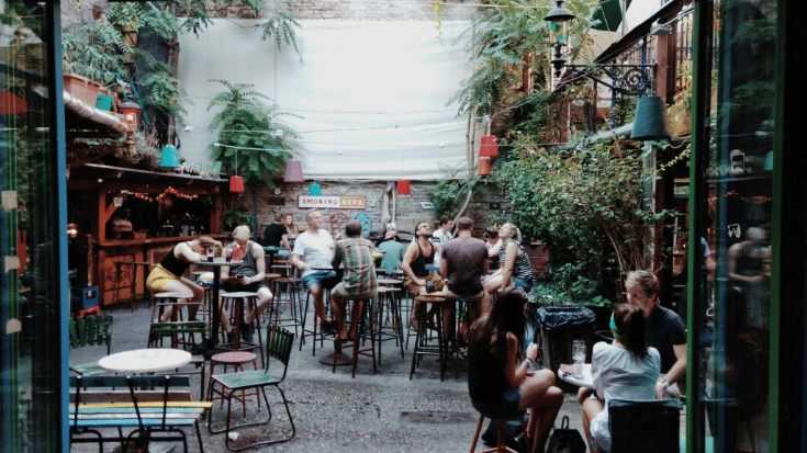 A photograph showing the courtyard of Budapest's Szimpla Kert ruin bar. There are groups of people at each table, both men and women, dressed in casual clothes and eating, drinking and socialising. To illustrate a blog post entitled 'The Ultimate Sziget Festival Guide.'