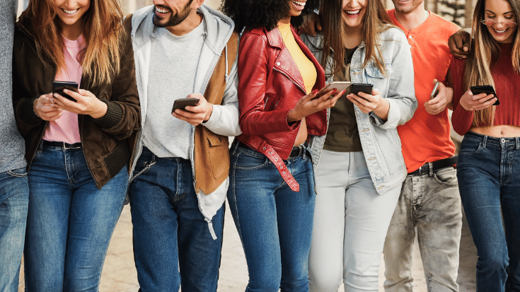 A colour photograph showing the mid-to-bottom-halves of a big group of people, probably friends, as they all check their phones and smile and laugh. To illustrate a blog post entitled 'The best group travel apps for 2025.'