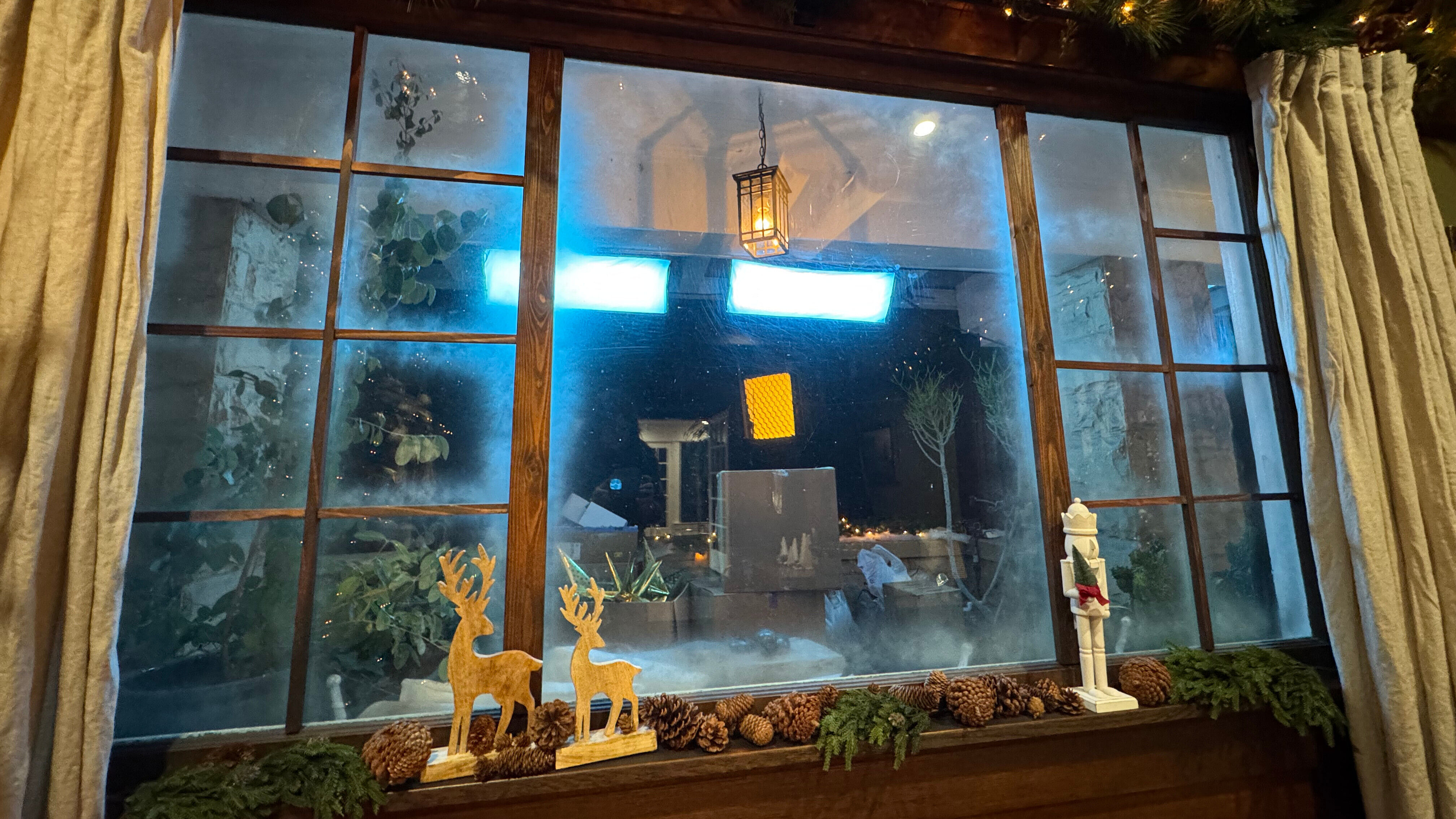 A living room window decorate for Christmas, complete with fake snow. Beyond the window, studio lights and empty boxes are visible.