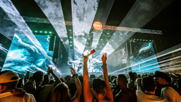 A photograph of a stage at a music concert at night. The stage is in the background, is lit by lasers and spotlights and has a sign above it saying 'Sziget'. In the foreground, the crowd is pictured and some people are holding their hands int he air, enjoying the music. To illustrate a blog post entitled 'The Ultimate Sziget Festival Guide.'
