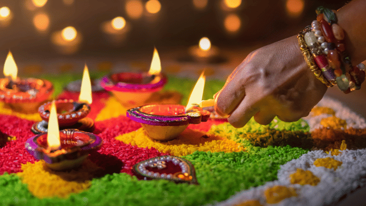 A photograph of close-up of an Indian woman's hand as she lights traditional candles called 'Diya lamps' during Diwali celebrations. The lights are small oval-shaped dishes decorated in bright colours such as reds, blues,pinks and yellows, and each has a small candle sitting in the middle. The lights are surrounded by brightly coloured flower petals in green, yellow and red and they have been arranged into a pattern. This image is to illustrate a blog post entitled When is Diwali 2025?'