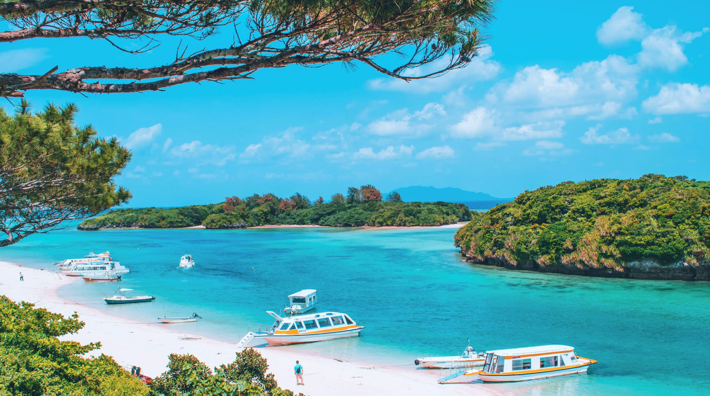 A colour photograph showing a white-sand beach with a clear azure blue sea. Sitting on the sea shore are three small white passenger boats/yachts, with stripes of yellow and blue decorating their hulls. There are a few figures dressed in beach wear wandering around on the sands alone, taking in the beautiful views. To illustrate a blog post entitled 'Top Things to Do in Okinawa, Japan's Tropical Gem.'
