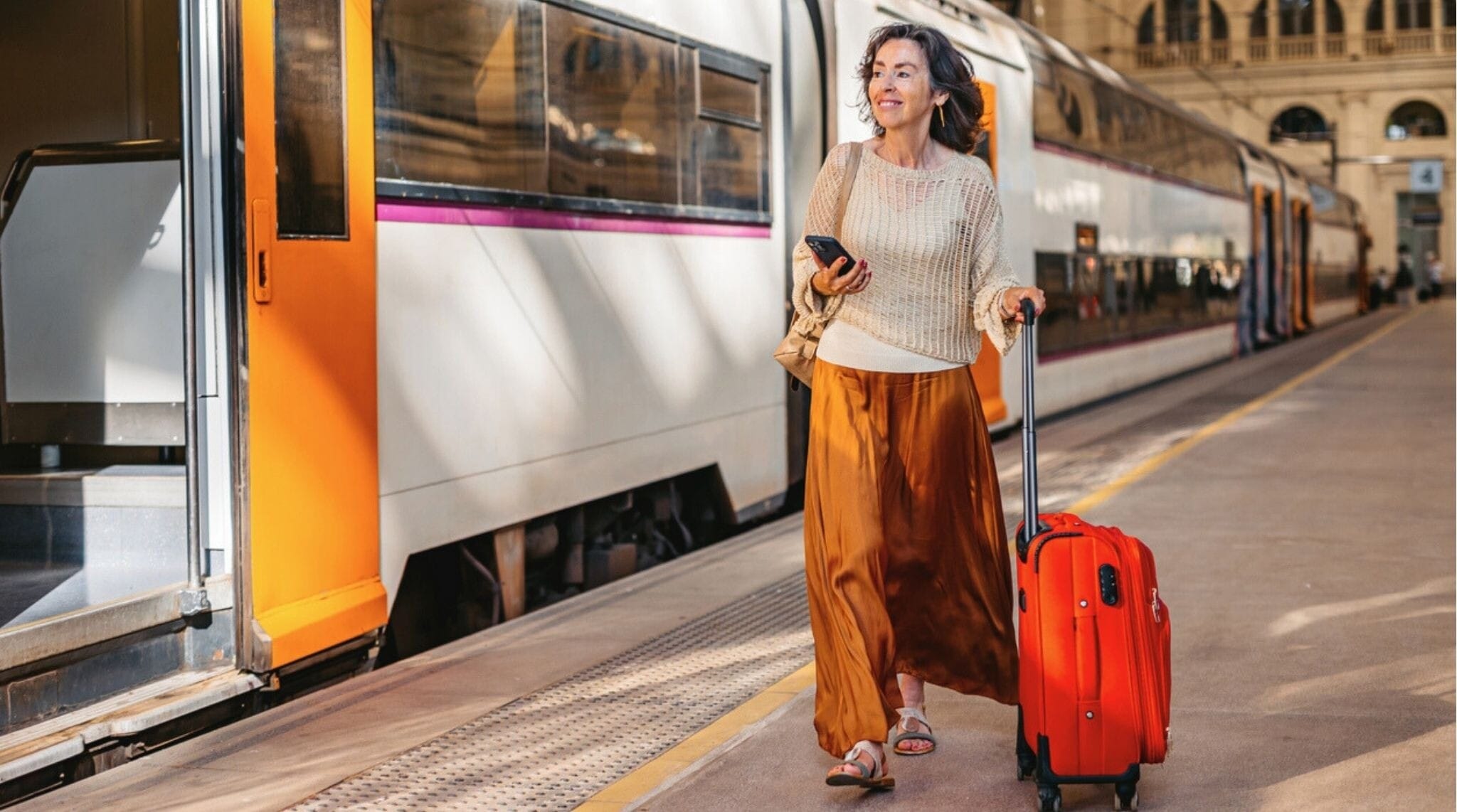 Viajera mayor consultando su teléfono con la maleta lista en una estación de tren europea.