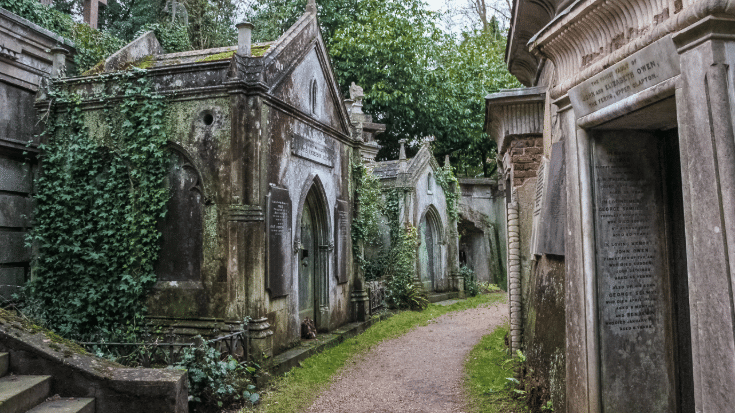 A colour photograph of Highgate Cemetery in London, UK. There is a grey gravel path winding to the right slightly and along the path are elaborately carved tombstones. Some are old and leaning over and stained by weather marks over time. The greenery is overgrown and includes ivy, bushes and trees. The graveyard has a Gothic and atmopsheric look to it. To illustrate a blog post entitled '10 of The World's Most Beautiful Cemeteries'.