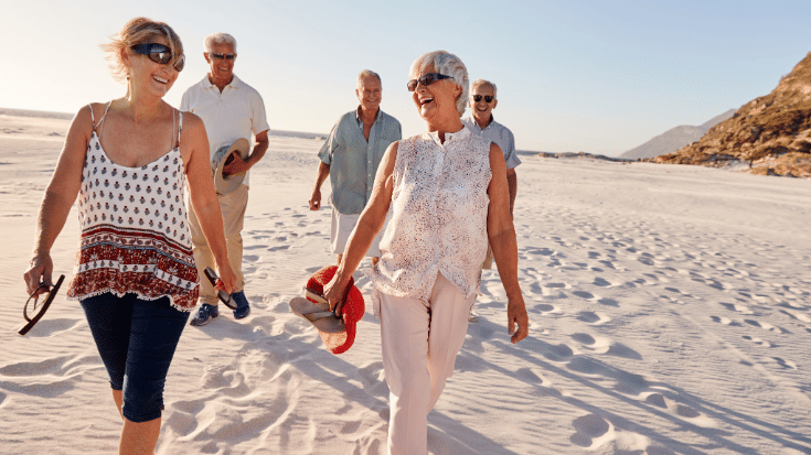 A colour photograph showing five people in their late fifties or early sixties walking on a beach together, talking and planning their travels. They're all smiling, wearing loose summer clothes and are walking barefoot across the sand. To illustrate a blog post entitled 'The best group travel apps for 2025.'
