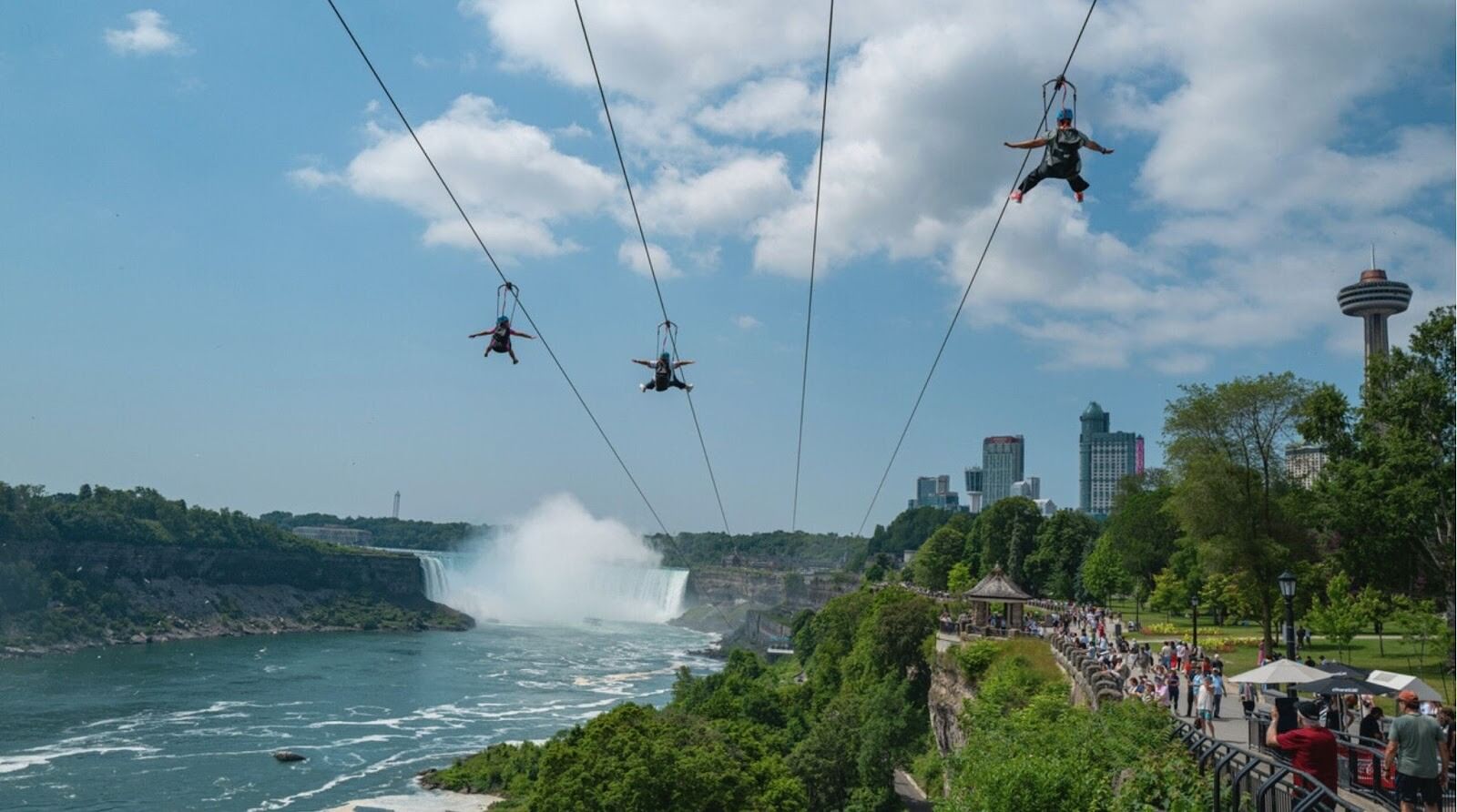 Três pessoas deslizam em tirolesas sobre a área do Niagara Parkway, com as Cataratas Horseshoe ao fundo.