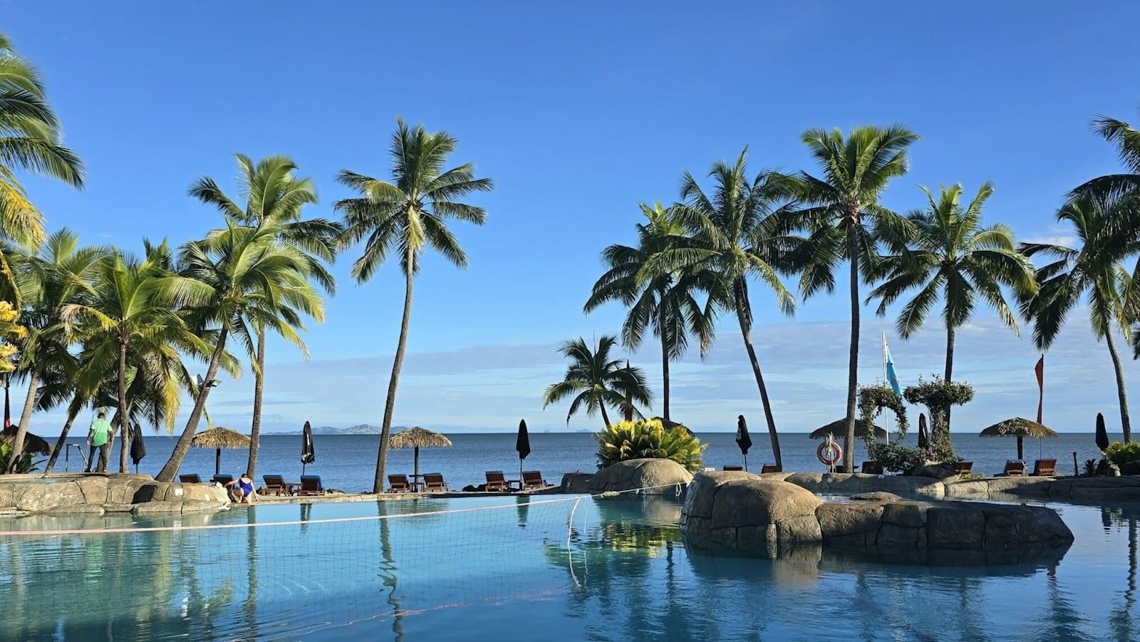 A large swimming pool, near a tropical ocean surrounded by tall palm trees