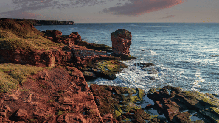 A color photograph showing the beautiful but choppy waters off the Scottish coast at Devil's Head in Arbroath, Scotland. It shows a towering red sandstone sea stack at Seaton Cliffs and colourful blue and pink sunrise. To illustrate a blog post entitled 'Guillermo Del Toro's Frankenstein Filming Locations.'