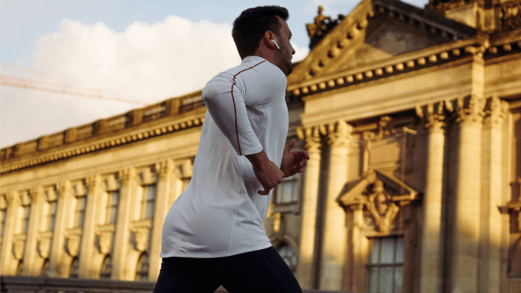 A colour photograph of a young man of asian descent with olive skin and black hair and beard dressed in a white running shirt and black running shorts. He is running past Berlin's famous Reichstag building. To illustrate a blog post entitled 'Berlin Half Marathon 2026: Can You Still Register?'