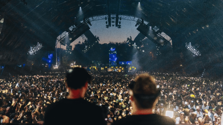 A photograph showing the blurred backs of two people's heads as they stand and perform on a large stage in the foreground. In the background, a large crowd has gathered and is listening to the music. To illustrate a blog post entitled 'The Ultimate Sziget Festival Guide.'