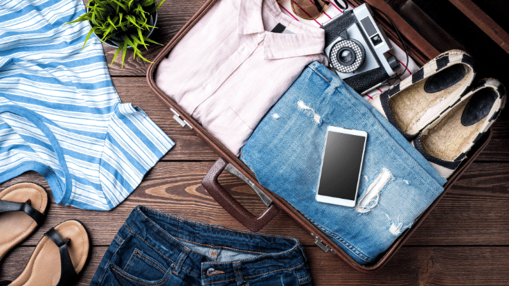 A photograph of an open hardshell suitcase on a wooden floor. The suitcase is almost completely packed with summer clothes such as sandals, shirts and denim shorts. There is also a black and white disposable camera sitting on top of the case and a white mobile phone sitting on the wooden floor next to the case. To illustrate a blog post entitled 'Where is Hot in November in Europe?'