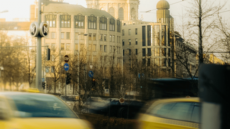 A photograph of Budapest city. In the foreground are blurred-out yellow taxis mid-journey. In the background are several grand looking buildings in a sandy beige colour. To illustrate a blog post entitled 'The Ultimate Sziget Festival Guide.'