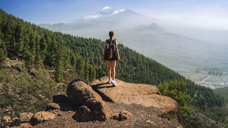 An image of a young woman hiking on the volcano Teide in Tenerife, Spain. You can see her from behind, with her backpack standing looking out over a blu sky with beautiful green evergreen trees all around her to illustrate a blog post entitled The Best Winter Family Holidays from Ireland.