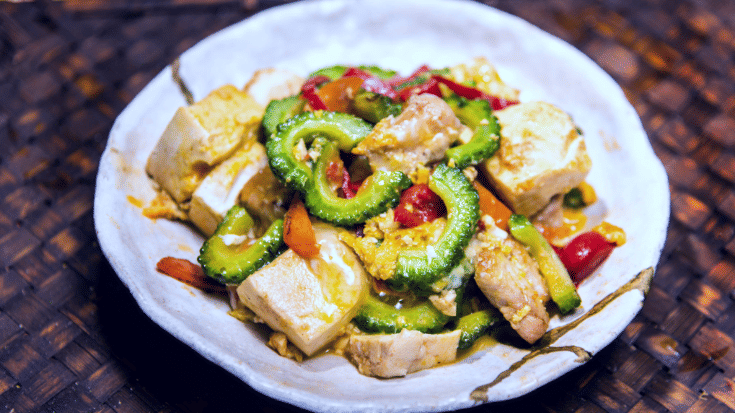 A colour photograph showing a close-up of one of Okinawa's most famous local dishes, goya chanpuru. On a bright white plate you can see cooked slices of the bitter melon plant goya, pork slices, egg, red peppers and tofu. To illustrate a blog post entitled 'Top Things to Do in Okinawa, Japan's Tropical Gem.'