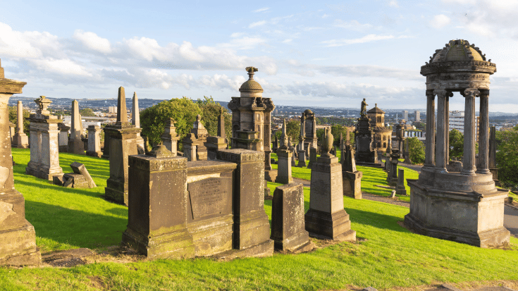 A colour photo of an historic Glasgow cemetery, the Necropolis, which sits on a hill outside of Glasgow Cathedral. In the image are elaborate and decorative grave sites in brown stone, marked by centuries of weather and wind. In the background the sky is blue with white clouds sitting across it. To illustrate a blog post entitled 'Guillermo Del Toro's Frankenstein Filming Locations.'
