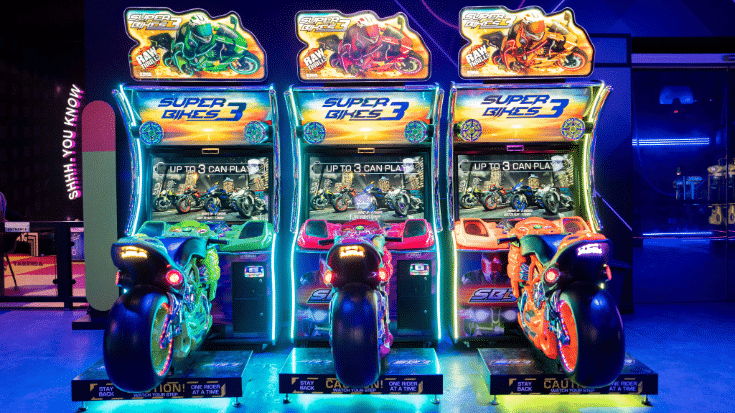 An image of three game arcade units at the Esports World Cup 2025, each has a large motorbike in front of a gaming screen and they are all lit in bright colours.