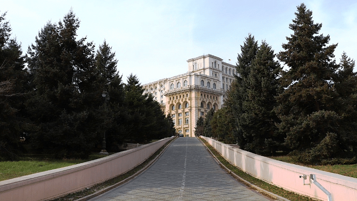 An image of the Palace of the Parliament on Bucharest, Romania, a pink and yellow building down a path flanked by fir trees, for a blog entitled Extreme Day Trips Abroad from the UK.