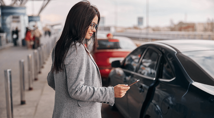 An image of a young woman with long brown hair, glasses and a grey suit jacket looking at her mobile telephone to book a taxi, while standing outside an airport terminal and holding her wheel along suitcase, to illustrate a blog post entitled the cheapest way to get to Heathrow Airport from London.