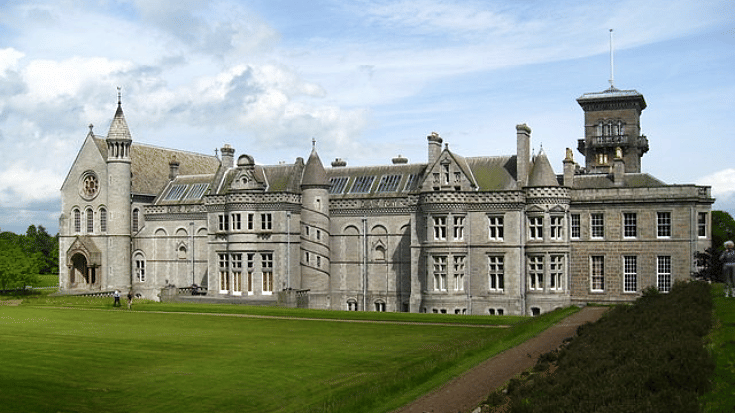 A colour photograph of Dunecht House, a stately home in Aberdeenshire, Scotland. It's made from grey stone and features round towers with pointed roofs, turrets and beautifully carved decorations and patterns on the outside of the building. In the foreground is a bright green, perfectly curated lawn and in the background you can see a clear blue sky dotted with white clouds. To illustrate a blog post entitled 'Guillermo Del Toro's Frankenstein Filming Locations.'