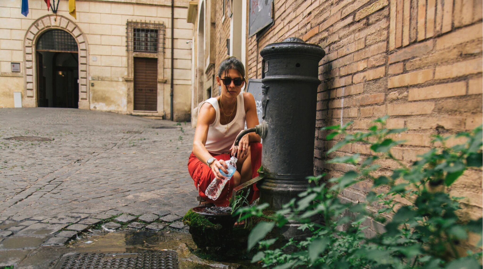 Uma mulher usando óculos de sol e roupas casuais coleta água limpa em uma garrafa plástica de uma fonte tradicional da cidade em um pitoresco beco de paralelepípedos cercado por arquitetura histórica europeia. Roma, Itália.