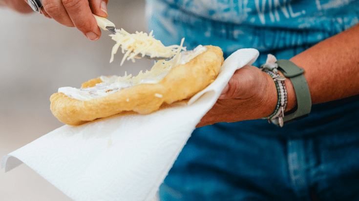 A photograph of a close-up of a woman's hand as she prepares the street food dish langos. It is a deep-fried crunchy pastry with a depression in the centre which is filled up with cream cheese before being topped with shredded cheese. To illustrate a blog post entitled 'The Ultimate Sziget Festival Guide.'