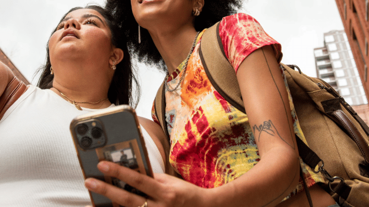 A photograph of two young women looking at a phone screen and speaking to one another in the foreground. One is wearing a white t-shirt and the other woman is wearing a warm-coloured tie dyed t-shirt. In the background are some red brick buildings in New York City. To illustrate a blog post entitled 'New York Comic Con 2025: A Complete Travel Guide for Attendees.'