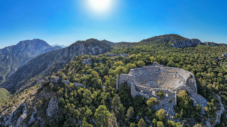 A photograph of Turkey's Taurus mountain range covered in lush greenery against a blue sky. In the foreground is the Termessos ancient city, showing the amphitheatre. Termessos is one of Antalya and Turkey's most outstanding archaeological sites. To illustrate a blog post entitled 'nine things to do in Antalya'.