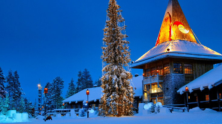 An image of a wooden house against a dark blue sky at twilight, with snow surrounding and a very tall Christmas tree with streetlights shining in the dark - this is Rovaniemi in Finnish Lapland said to be the Christmas capital of the world and home to Santa Claus himself, image to illustrate a blog post entitled The Best Family Winter Holidays from Ireland.