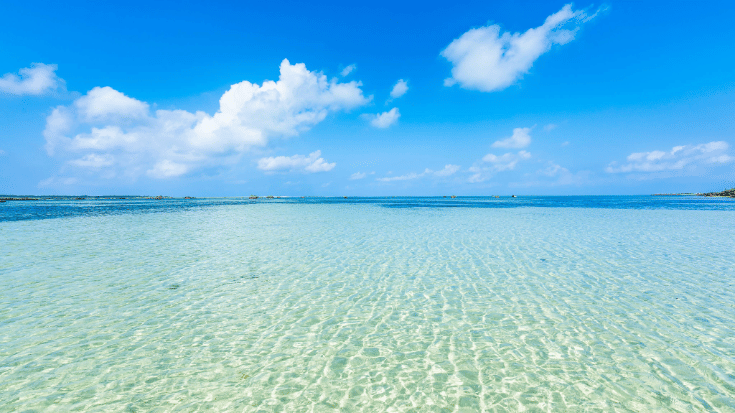 A colour photograph showing the shallow waters reflecting the bright sunshine on a white-sand beach on Miyako Island, Okinawa, Japan. In the foreground you can see the sand and the ocean water stretching out into the distance. overhead, the sky is bright blue with white clouds dotted through it. To illustrate a blog post entitled 'Top Things to Do in Okinawa, Japan's Tropical Gem.'