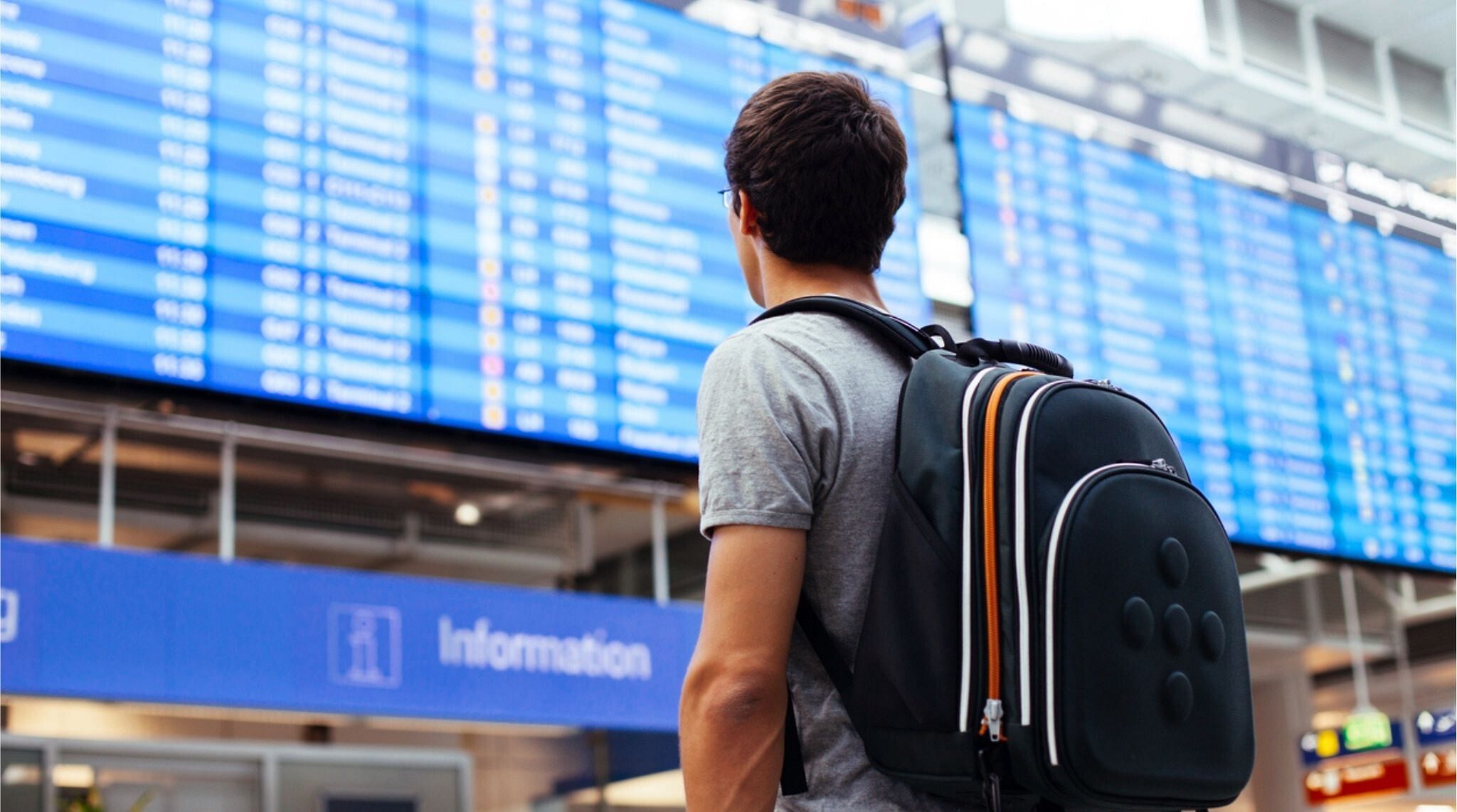 Rapaz olhando o painel de voos do aeroporto com uma mochila nas costas