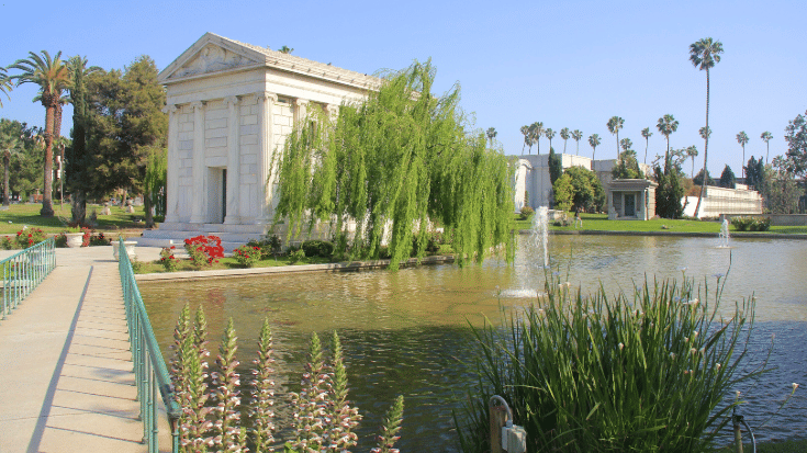 A colour photograph of Hollywood Forever Cemetery in Los Angeles, USA. In the background, there are palm trees dotted through the image, the sky is bright blue and it appears to be a sunny and hot summer day. There is a large Greek-style building with Corinthian columns and made of white stone in the background. It sits on a bright green, very well-kept lawn and is next to a large lake. Overhanging the lake is a yew tree, whose branches are blowing in the light breeze. To illustrate a blog post entitled '10 of The World's Most Beautiful Cemeteries'.