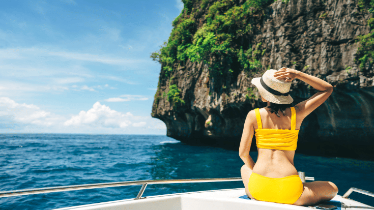 An image of a woman in a yellow two-piece swim suit from behind, cross-legged on a small boat, holding on to a sun hat she is wearing. In the background is a Thai cliff face and the bright blue Thai waters and sky - an image to illustrate a blog post entitled '33 fun facts about Thailand'. 