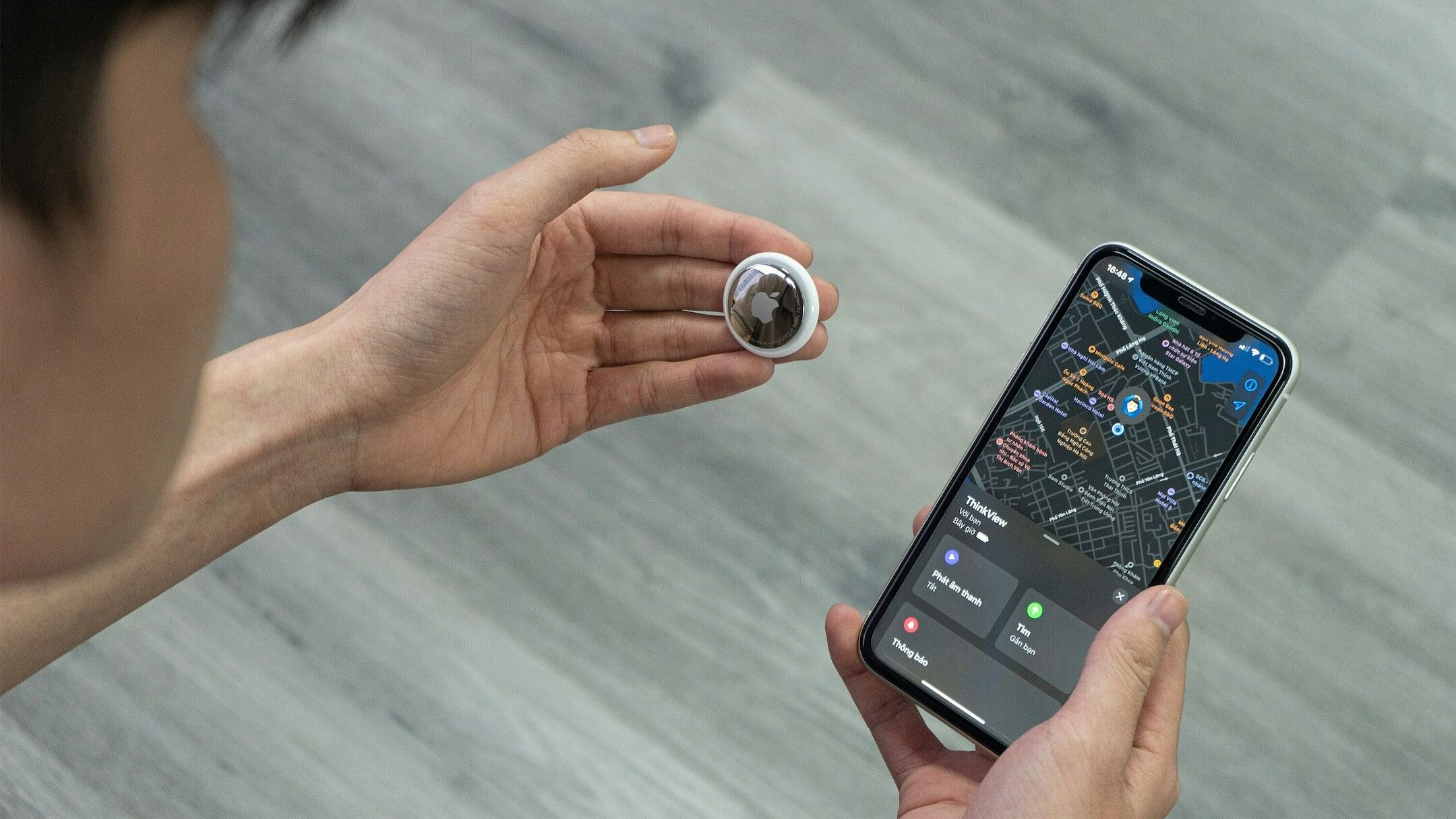 A man holds an Apple AirTag in one hand with his iPhone in another. The phone screen shows a map.