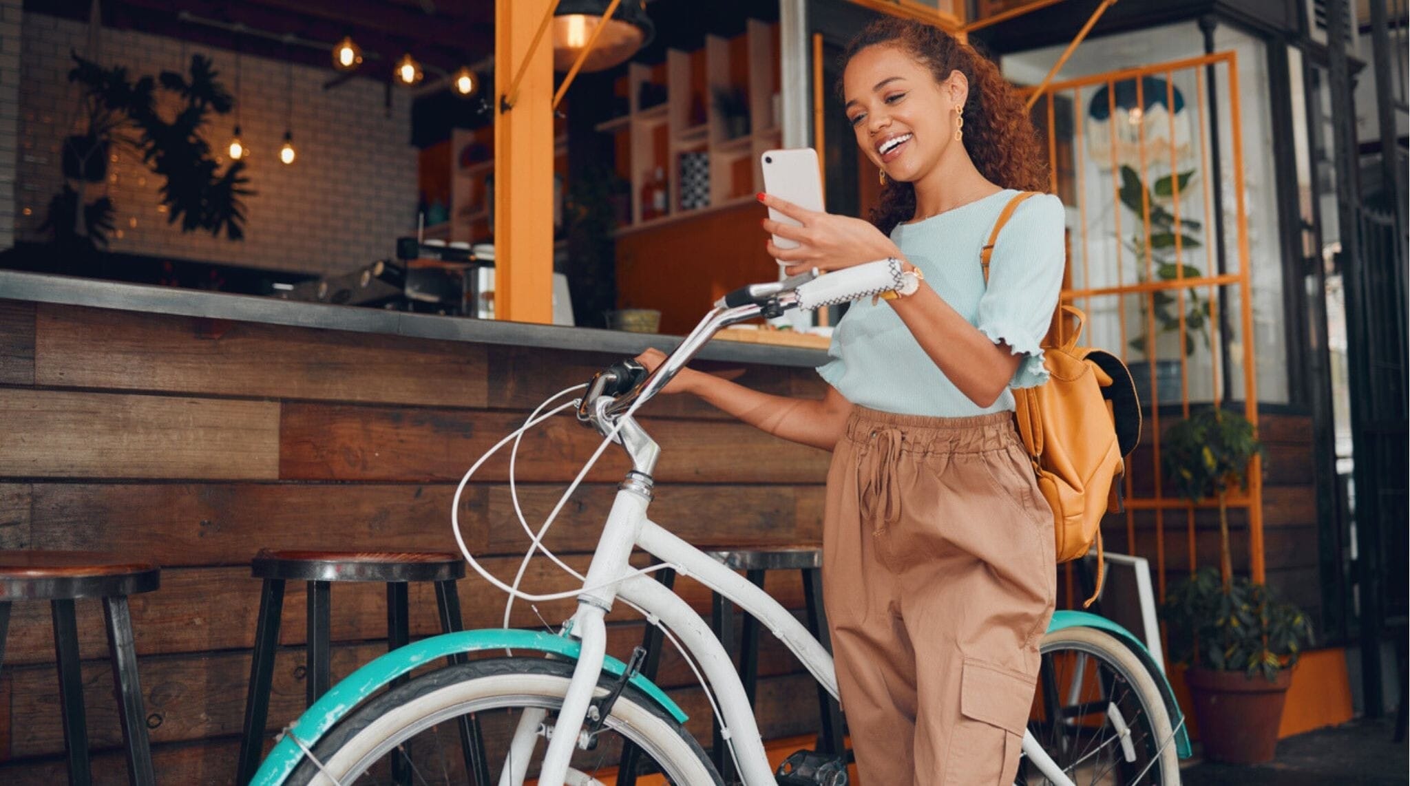 Mujer usando su teléfono en una cafetería urbana durante un viaje, con bici y mochila de día