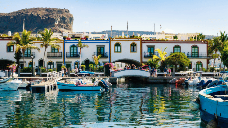 A photograph of Puerto de Morgan on Gran Canaria Island, Spain. In the foreground is a body of blue water and int he background is a row of whitewashed houses, with blue and brown trims painted around their windows and doors. There are people dressed in summer clothes walking in front of the houses and enjoying the views across the water. There are also green leaved palm trees at the front of some of the houses and the sky is a very clear blue. To illustrate a blog post entitled 'Where Is Hot in November in Europe?'