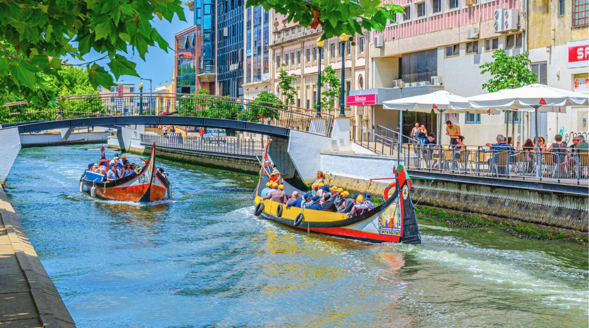 Aveiro, Portugal, 13 de junho de 2017: Barco tradicional colorido moliceiro com turistas navegando por um canal estreito, com ponte sobre o curso d’água e edifícios ao longo do calçadão na área central da cidade.